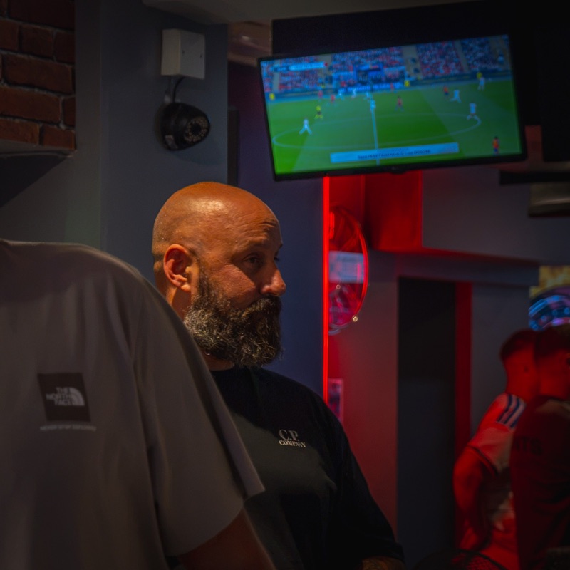 A man with a grey beard at the bar, a football match on the screen above him