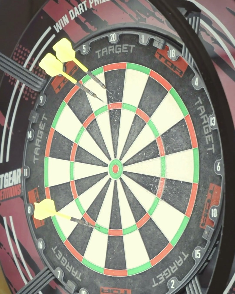 A dartboard with three yellow-flighted darts in it, “Win dart prizes” lettered round its surround