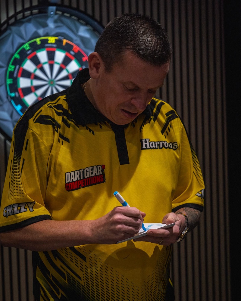 Dave Chisnall in his yellow Dart Gear Competitions shirt, signing a card in front of a dartboard