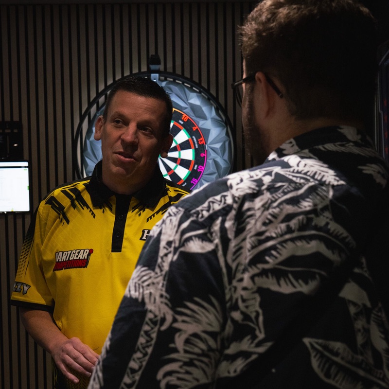Dave Chisnall in his yellow Dart Gear Competitions shirt, talking with a visitor in front of a dartboard