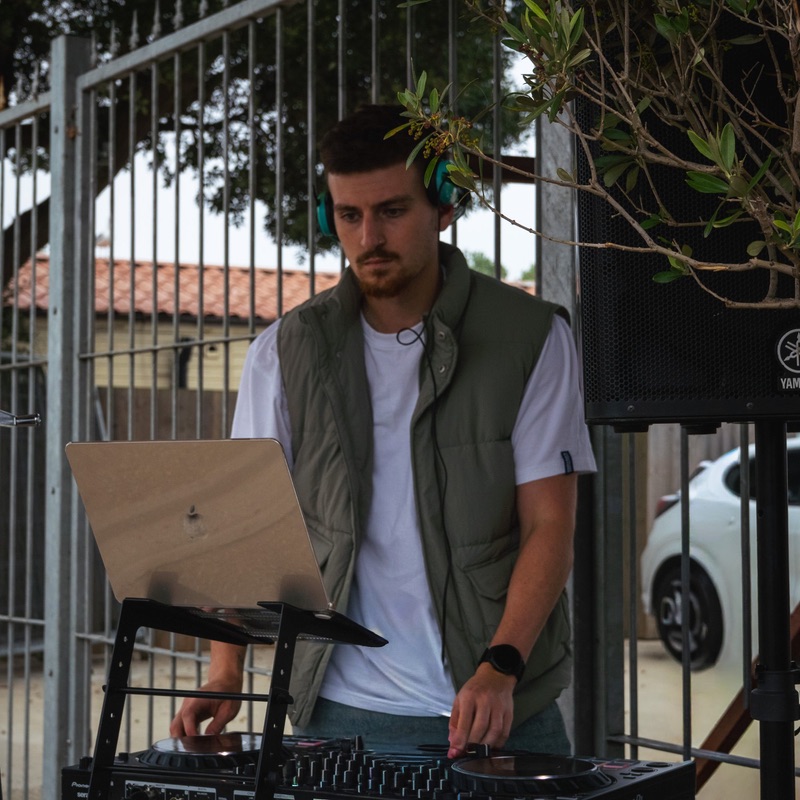 A DJ in a green gilet and headphones at the decks outdoors, a laptop on a stand beside him