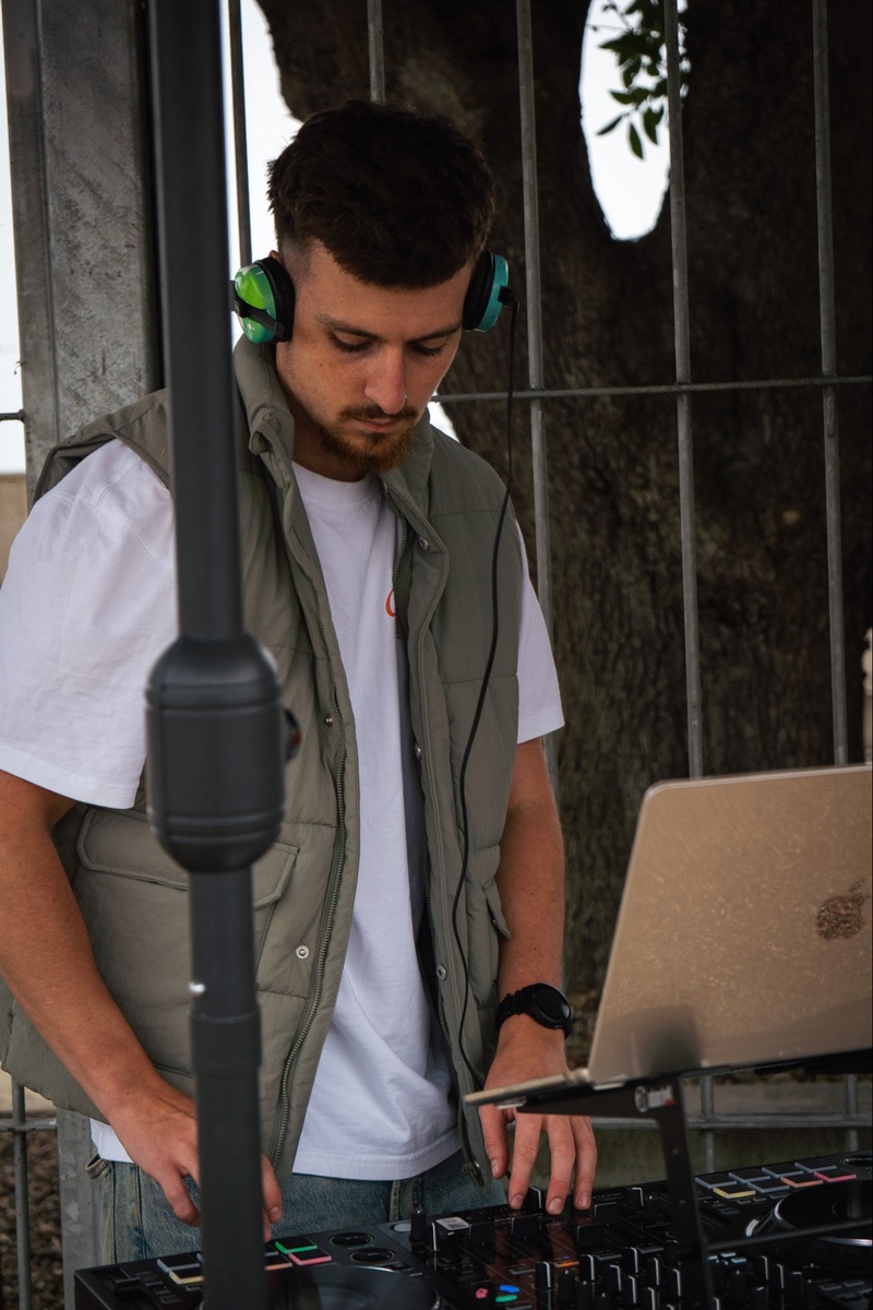 The DJ looking down at the decks with his headphones on, seen past a speaker stand