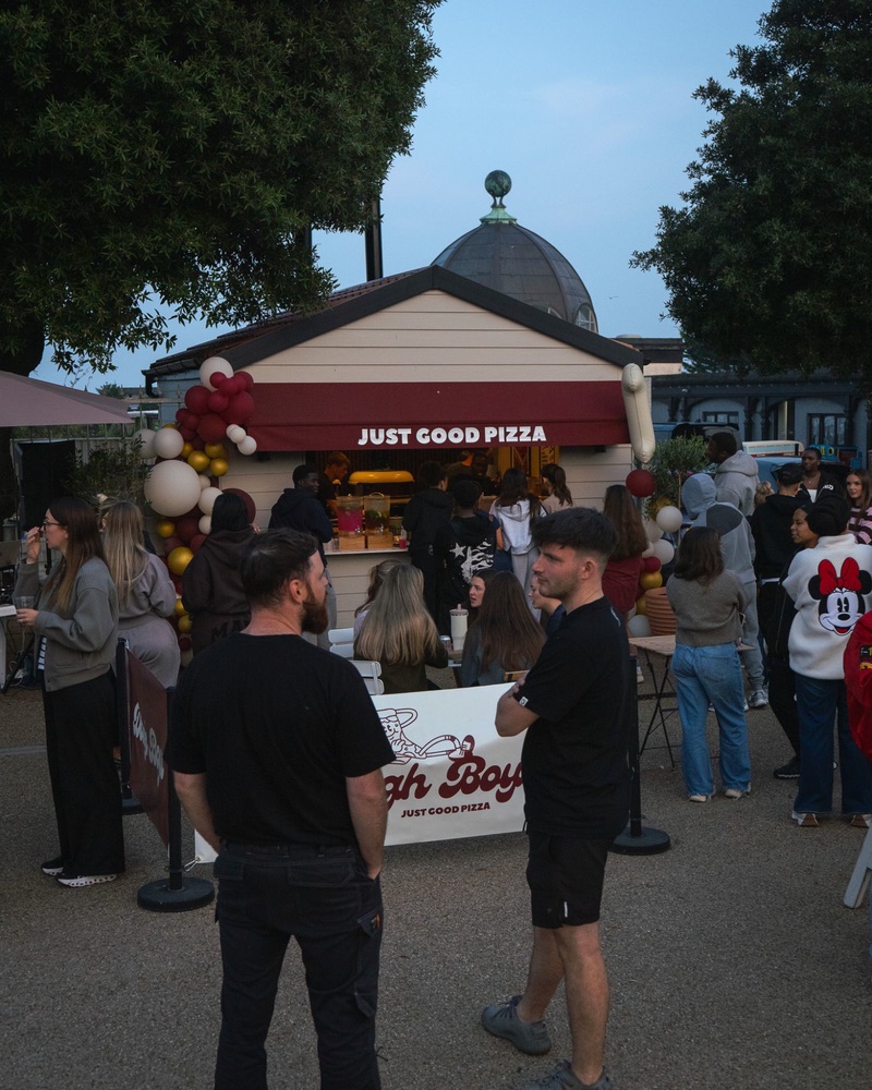 A crowd outside the Dough Boys kiosk at dusk, balloons by the hatch and a Dough Boys banner on the barrier