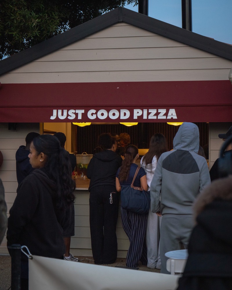 People queueing at the serving hatch under the red awning lettered Just Good Pizza
