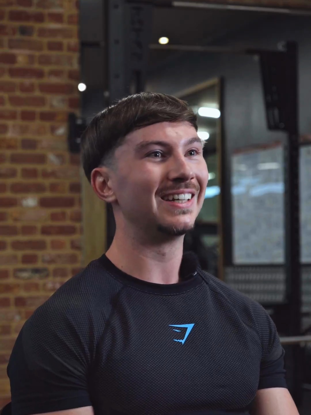 Harrison, a personal trainer in a black training top, laughing mid-interview in front of a brick wall